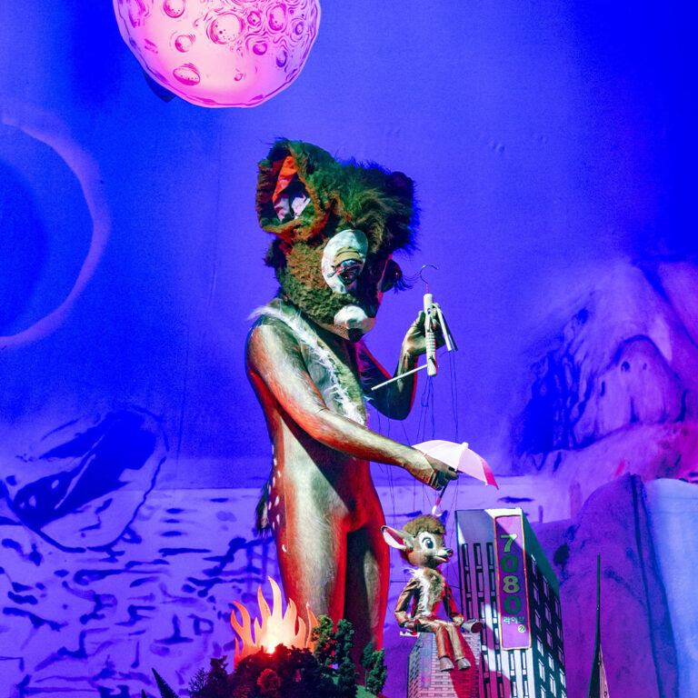 Eine Person in einem Rehkostüm mit einem flauschigen Rehkopf steht in einer violetten Landschaft. In den Händen hält die Person eine Bambi-Reh-Marionette. Im Vordergrund sind brennende Bäume zu erkennen und eine Stadtlandschaft ist angedeutet. Darüber scheint der Mond.